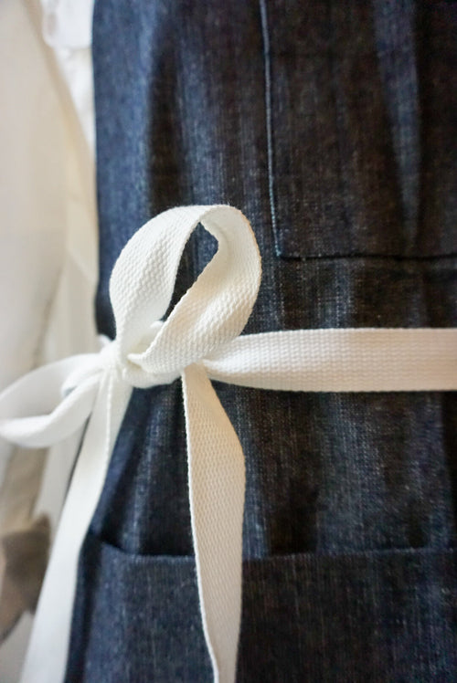 Close-up of a white ribbon tied around a dark denim apron (TANN Schürze).
